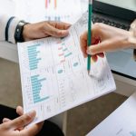 Woman pointing to a paper with a pencil that is held by another person that has graphic representations of statistics.